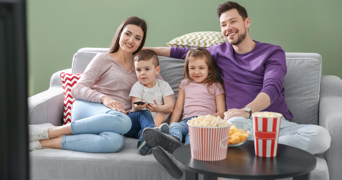Una familia sentada en el sillón viendo televisión junta, con las niñas y los niños acompañados por sus adultos